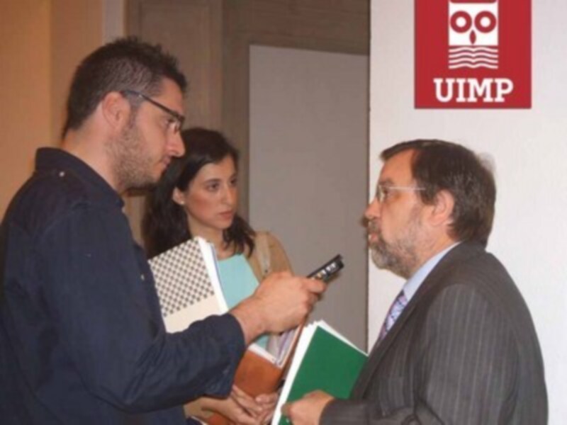 Dos personas en una reunión, uno sostiene un teléfono móvil y el otro parece estar hablando. En la pared hay un logo de UIMP.