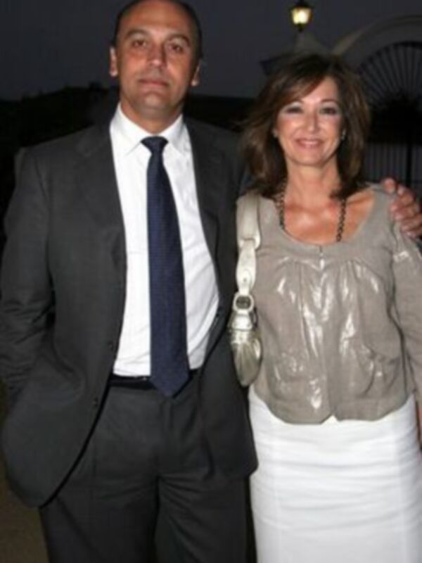 Una pareja posa juntos en una noche de eventos, el hombre lleva traje y corbata, la mujer usa vestido blanco y bolso.