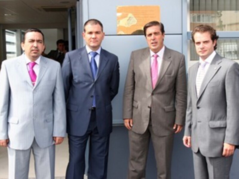 Cuatro hombres en trajes formales posan juntos frente a una puerta con un letrero en la fachada de un edificio.