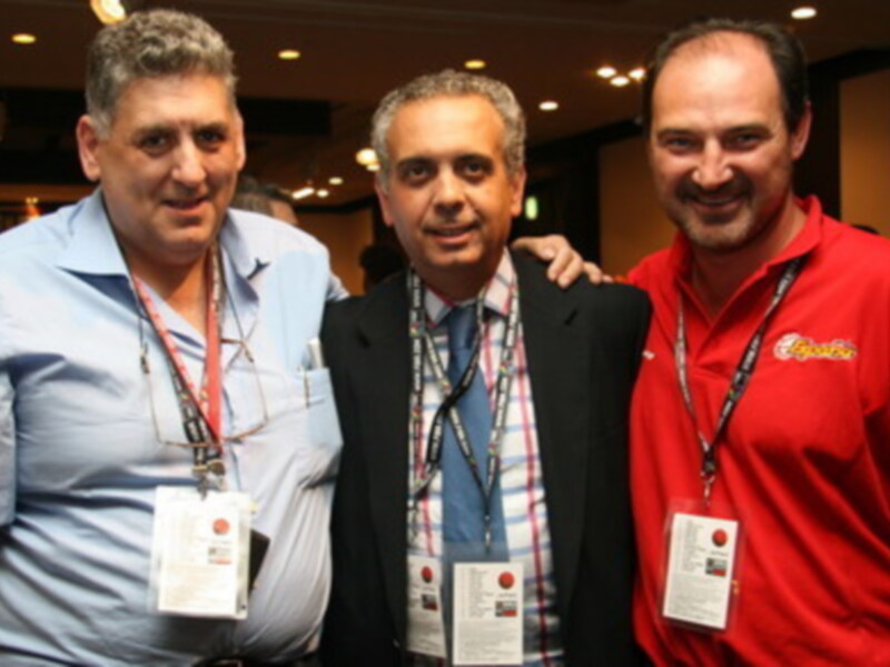 Tres hombres sonrientes posando juntos en un evento, con identificaciones de conferencia colgadas al cuello.