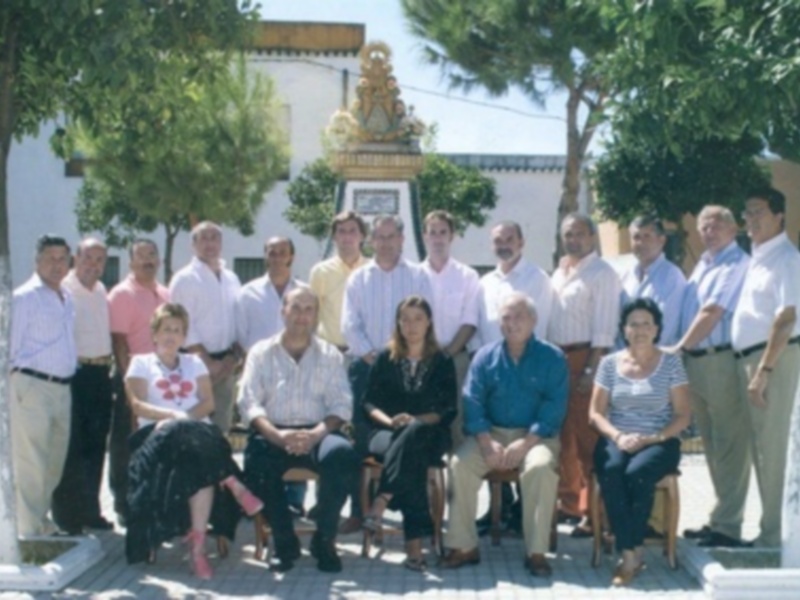 Grande grupo de personas reunidas al aire libre, posando para una foto familiar en un entorno soleado con árboles y edificios blancos en el fondo.