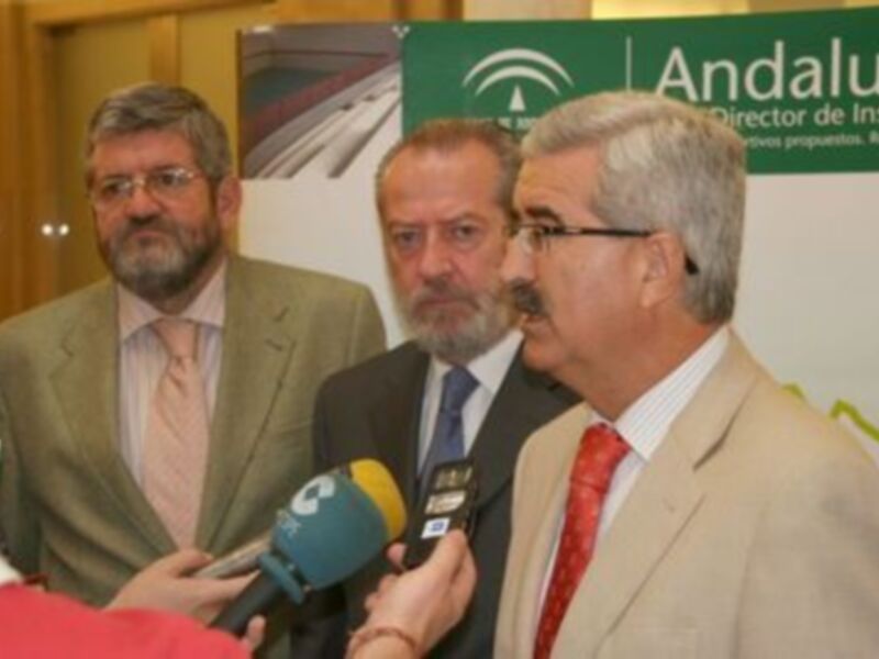 Tres hombres en trajes, uno de ellos siendo entrevistado por un micrófono, con un fondo que incluye una imagen de una estación de tren y un letrero de Andalucía.