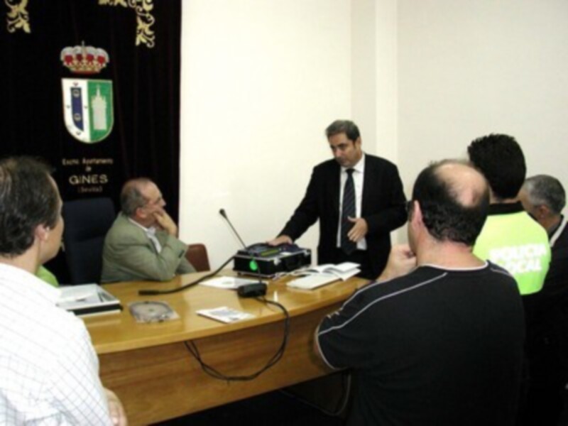 Persona hablando en una reunión o conferencia, con un escudo de armas en el fondo y un grupo de asistentes sentados en la mesa.