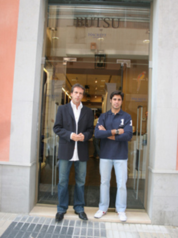 Dos hombres posando frente a una tienda de ropa, con el nombre 'BUTSU' en la puerta.