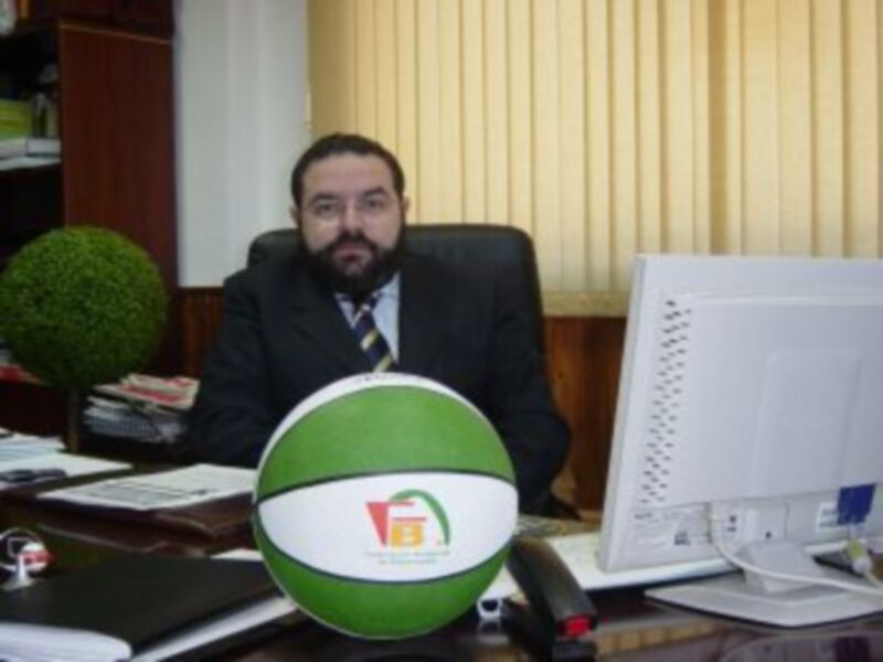 Un hombre en traje y corbata se sienta detrás de una mesa con un balón de baloncesto verde y blanco en primer plano, en un entorno de oficina.