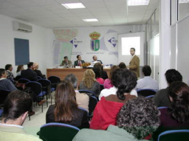 Un evento de conferencia en una sala con una pantalla proyectando información, con asistentes sentados en filas y un hombre en el centro del escenario. La sala tiene una decoración con un escudo y un logo en el fondo.