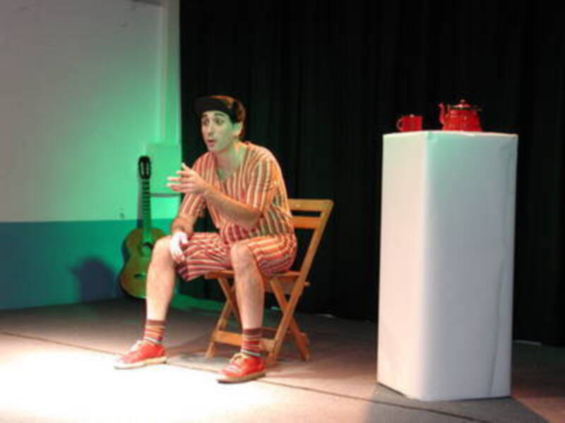 Actuación teatral con un personaje en traje de payaso sentado en una silla, con un pedestal y una guitarra al fondo.