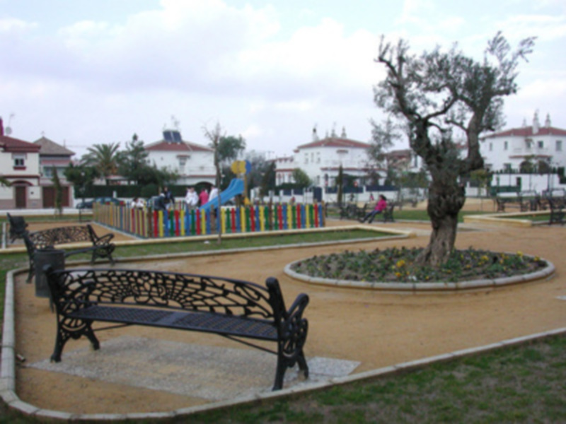 Parque urbano con bancos, árboles, y área de juegos infantil bajo un cielo parcialmente nublado.