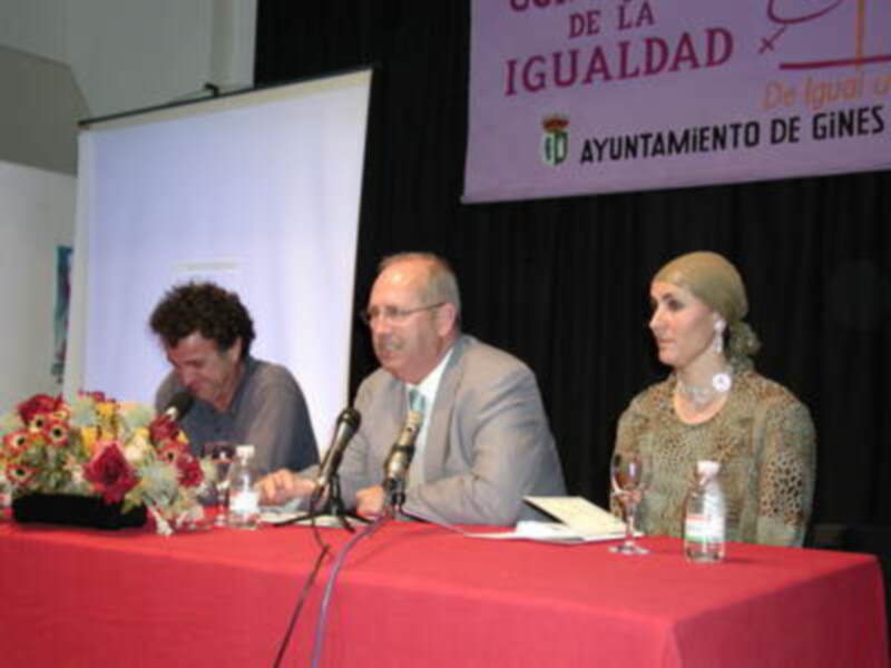 Tres personas sentadas en una mesa con micrófonos, en un evento del Ayuntamiento de Gines sobre la igualdad.