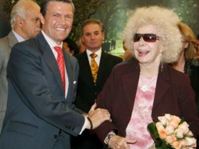 Un hombre en traje azul y corbata roja sonríe mientras sostiene la mano de una mujer mayor con una chaqueta marrón y un ramo de flores. Detrás de ellos, otros individuos observan la escena.
