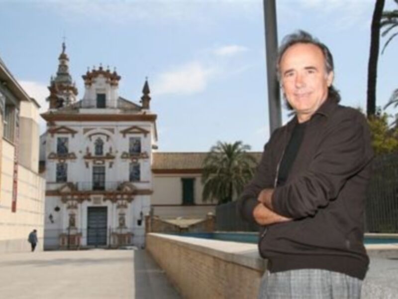 Un hombre posa frente a una iglesia histórica en un entorno urbano con palmeras y edificios modernos al fondo.