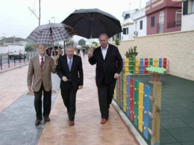 Tres hombres caminando bajo paraguas en un entorno urbano con un patio infantil y edificios al fondo.