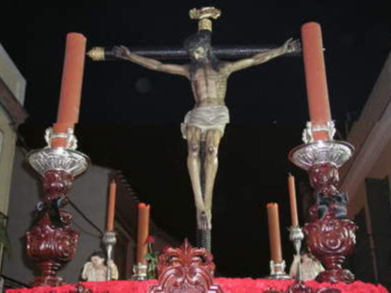 Procesión religiosa con estatua del Cristo de la Buena Muerte en la noche, flanqueada por candelabros altos.