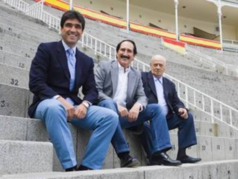 Tres hombres sentados en las gradas de un estadio, con banderas de España en el fondo.