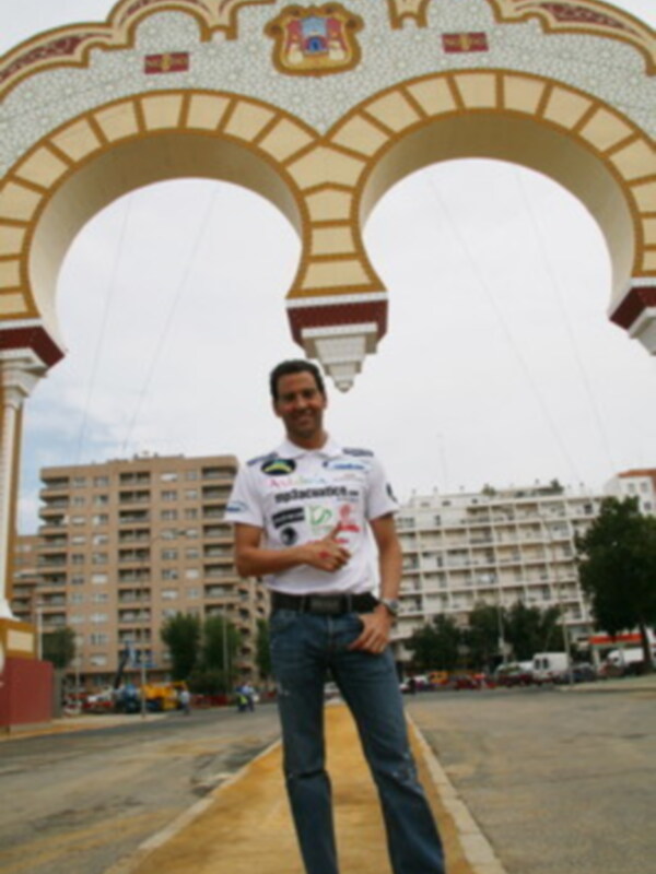 Persona posando frente a un arco decorativo en una plaza urbana, con edificios al fondo.