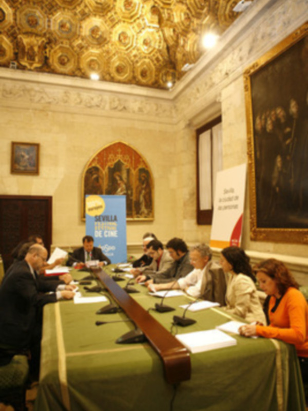 Una reunión formal en un salón con techo dorado, personas sentadas en una mesa, y una pancarta azul con texto en el centro.