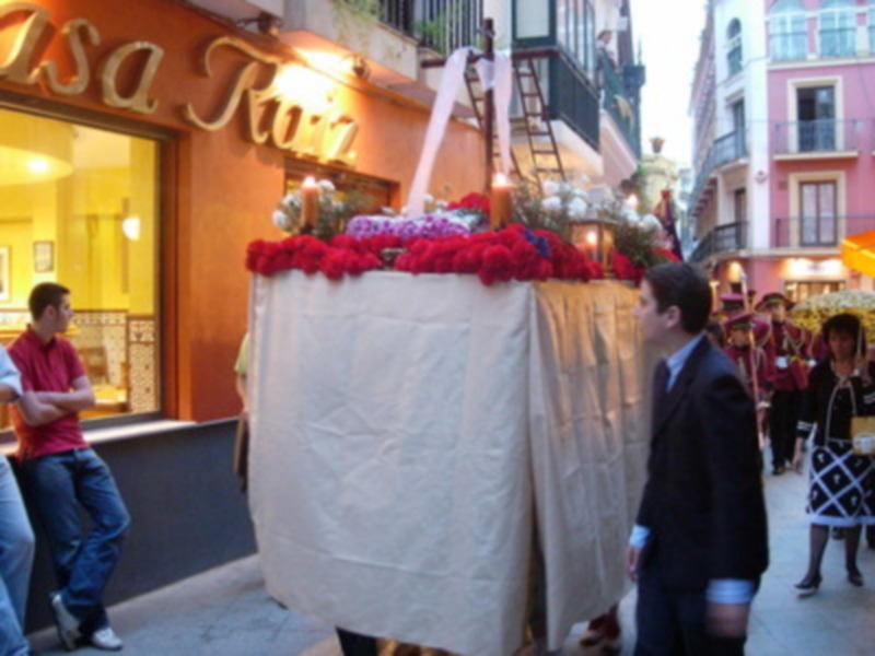 Procesión de Semana Santa con pasos procesionales y personas participando en la celebración en una calle de España.