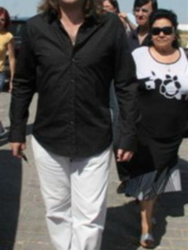 Persona caminando en una calle con otras personas al fondo, vistiendo una camisa negra y pantalones blancos.
