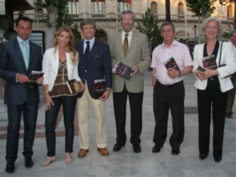 Una reunión formal con cinco personas posando para una foto, todos vestidos con trajes y pantalones, sosteniendo libros. El ambiente parece ser un evento o presentación de libros en una ubicación al aire libre, posiblemente en un parque o plaza.