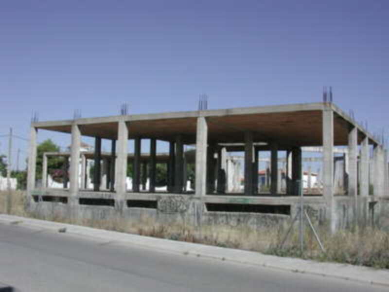 Estructura de hormigón en construcción con vigas de acero en la parte superior, ubicada en un entorno urbano con vegetación y una carretera a la izquierda.