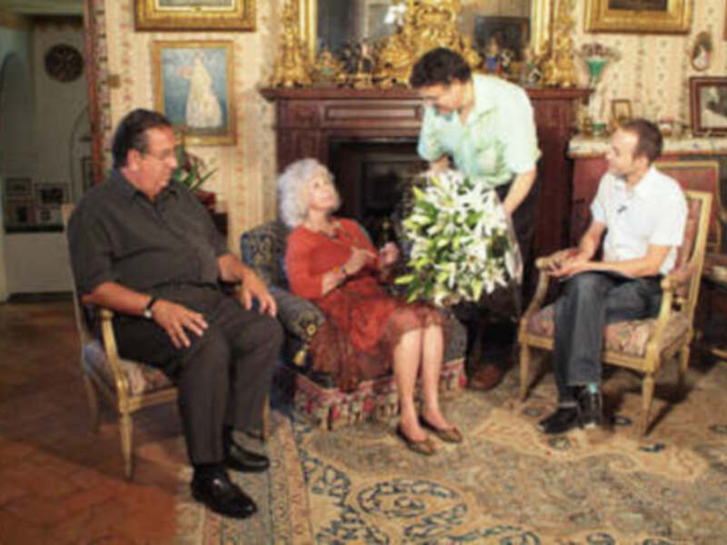 Una escena familiar en un entorno elegante, con una mujer en el centro, sosteniendo un ramo de flores, rodeada por dos hombres sentados en sillones, y una mujer en el fondo, todo en un ambiente de lujo con chimenea y cuadros.