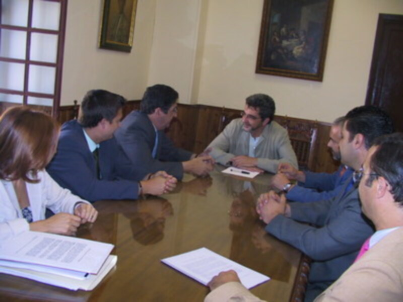 Una reunión formal en una sala con dos grandes ventanas, donde cinco personas están sentadas alrededor de una mesa, discutiendo y sosteniendo documentos.