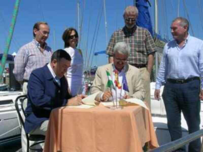 Un hombre firma documentos en una ceremonia al aire libre con veleros en el fondo.