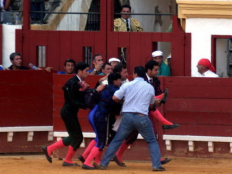 Un torero en el centro de la imagen, rodeado por varios hombres en trajes de charro, con una torera en el fondo. La imagen parece ser de una corrida de toros en un anfiteatro tradicional.