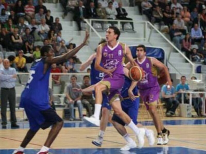 Jugadores de baloncesto en acción durante un partido, con un jugador en amarillo controlando el balón mientras otros luchan por la posesión en un entorno deportivo con público en el fondo.
