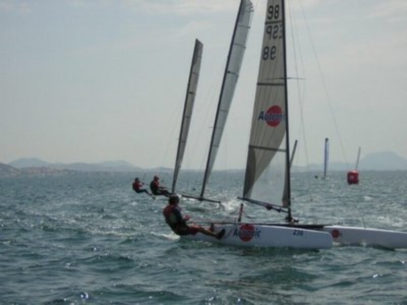 Tres personas en una embarcación de vela en el mar, con una señal de seguridad en el fondo.