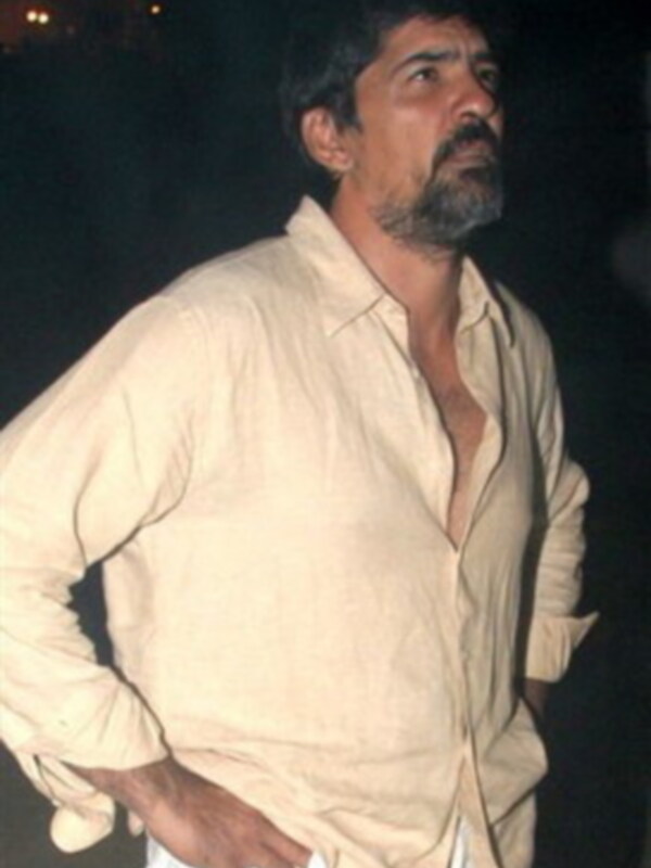 Hombre con camisa blanca abierta, posando con una mano en la cadera en un entorno nocturno.