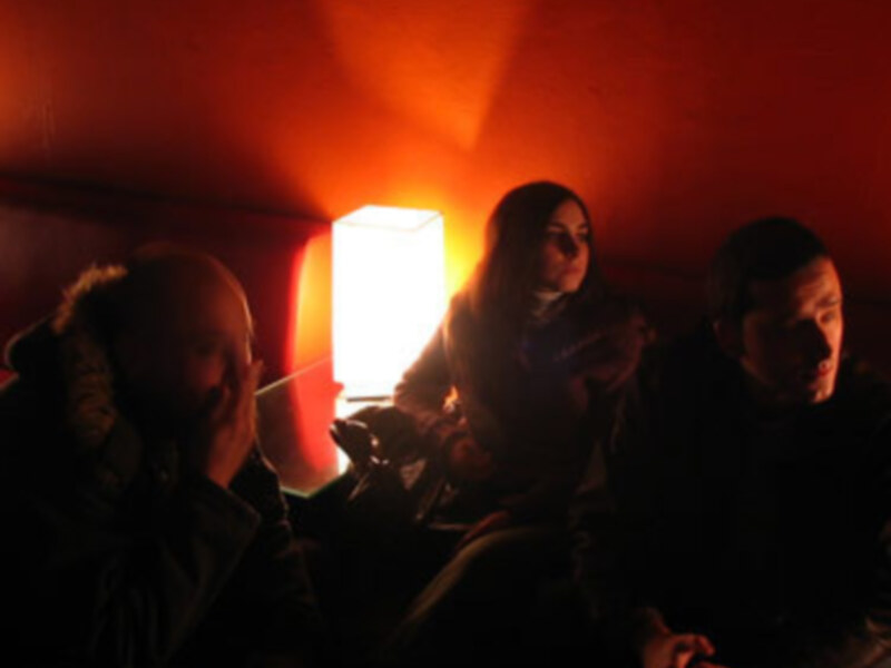 Tres personas sentadas en una habitación oscura, con una luz roja en el fondo. La atmósfera parece ser de una reunión o conversación en privado.