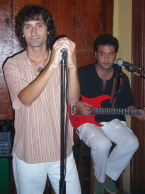 Dos músicos en vivo, uno tocando la guitarra y el otro cantando con micrófono en mano.