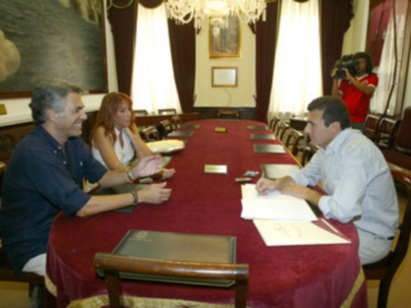 Una reunión formal en una sala con muebles clásicos y una mesa cubierta con un mantel rojo. Dos personas se sientan en la mesa, mientras que una tercera persona toma una foto. La sala tiene una gran pintura en el fondo y una lámpara de techo.