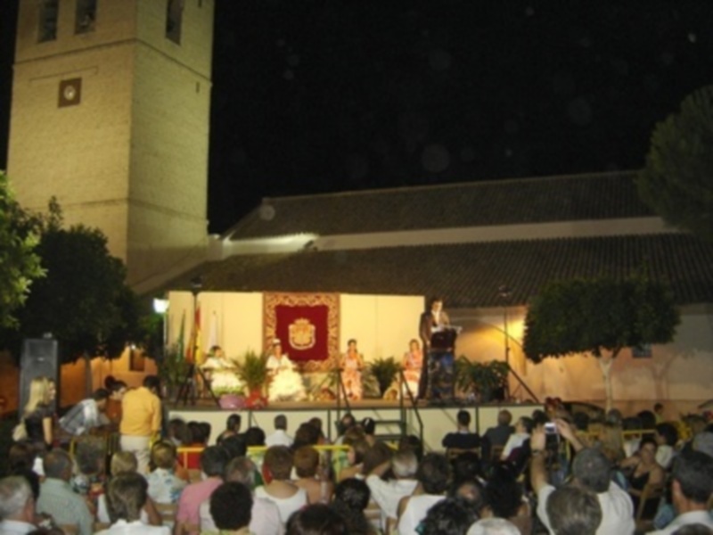 Escenario al aire libre con una banda y público en una noche de verano, frente a una iglesia iluminada.