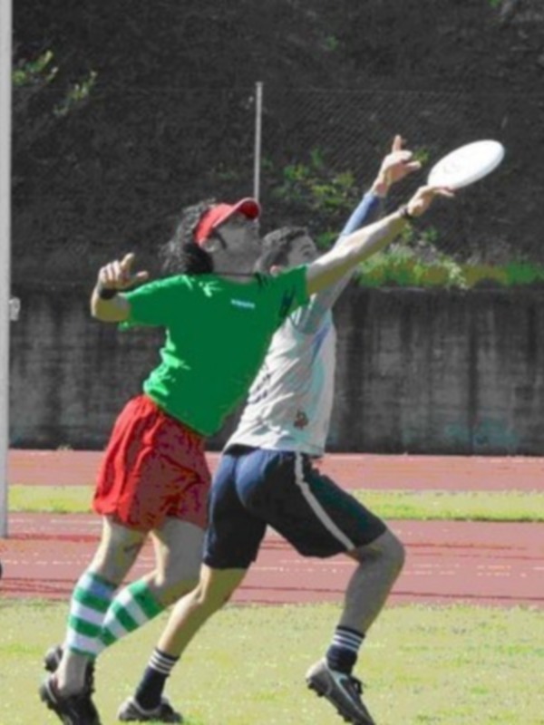 Dos personas jugando con un disco en un campo deportivo, uno con una camiseta verde y otro con una camiseta blanca y pantalones cortos azules, ambos con calcetines de colores diferentes. El campo tiene una pista de atletismo al fondo y árboles alrededor.