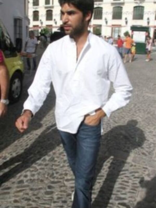Un hombre con una camisa blanca y pantalones vaqueros caminando en una calle con piedras. Detrás de él, hay edificios con ventanas y una persona caminando en la distancia.