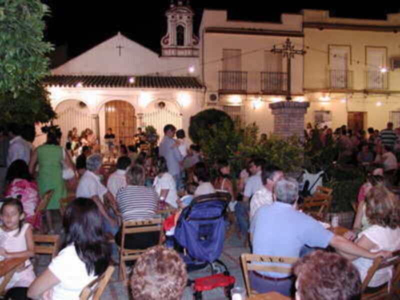 Fiesta nocturna en una plaza con gente sentada en mesas al aire libre, iluminada por luces y rodeada de edificios históricos.