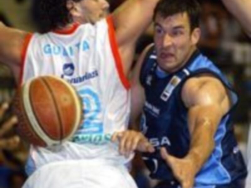 Jugadores en acción durante un partido de baloncesto, uno en blanco y otro en azul, mostrando intensidad y competencia.