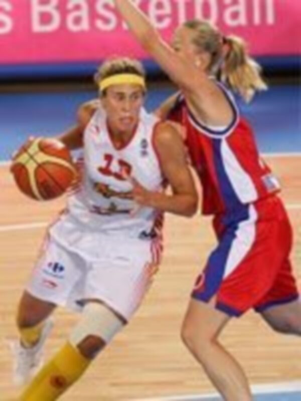 Una jugadora de baloncesto en acción, con el balón en la mano, mientras su compañera de equipo la observa.