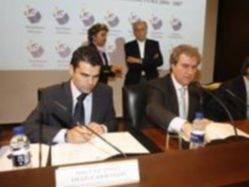 Conferencia de prensa con dos hombres sentados en una mesa con micrófonos y documentos, en un ambiente formal.