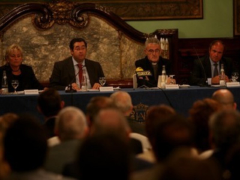Una reunión formal con personas sentadas en una mesa, probablemente en una sala de conferencias o una sala de audiencias, con un fondo decorado y una audiencia de personas en el frente observando.