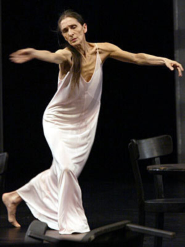 Danza clásica en movimiento fluido, vestida con un vestido largo blanco en un escenario oscuro.