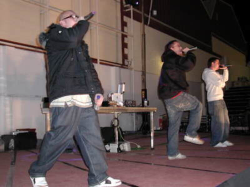 Tres personas cantando en un escenario con micros en la mano. La escena parece ser una actuación musical en un evento nocturno. El ambiente sugiere una atmósfera de entretenimiento y entretenimiento musical.