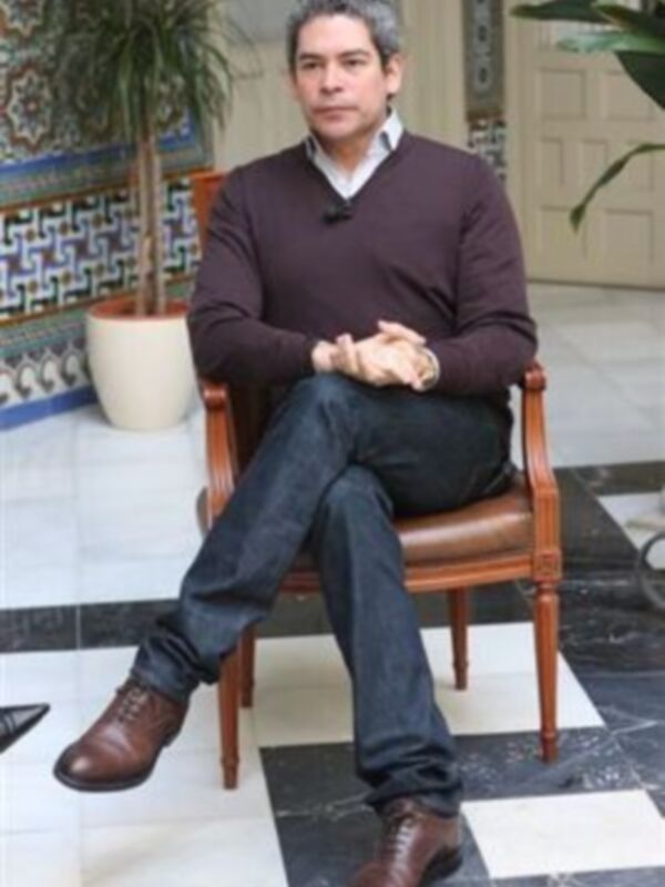 Un hombre sentado en una silla de cuero, con una camisa blanca y un suéter marrón, posando para la cámara. Fondo con azulejos de diseño y una planta en el lado izquierdo.