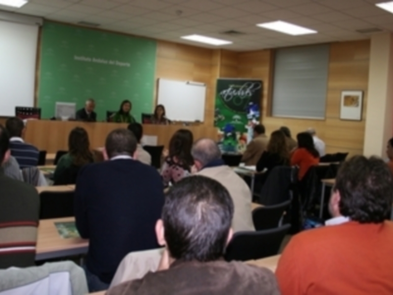 Una sala de conferencias con una pantalla verde en el fondo, personas sentadas en el público y un hombre en el escenario.