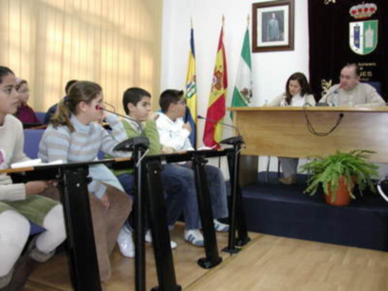 Reunión de estudiantes y autoridades en una sala de conferencias con banderas y un panel oficial en el fondo.