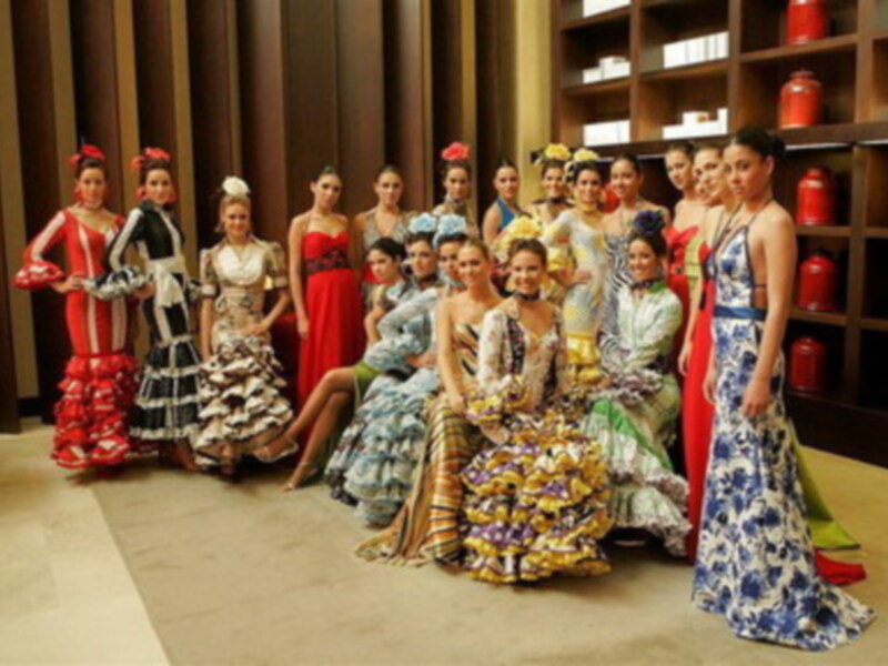 Una fila de mujeres vestidas con trajes de flamenco posando para una foto. Los vestidos son de colores vibrantes y variados, con detalles en flores y encajes. Al fondo, se pueden ver estanterías con objetos rojos y blancos.