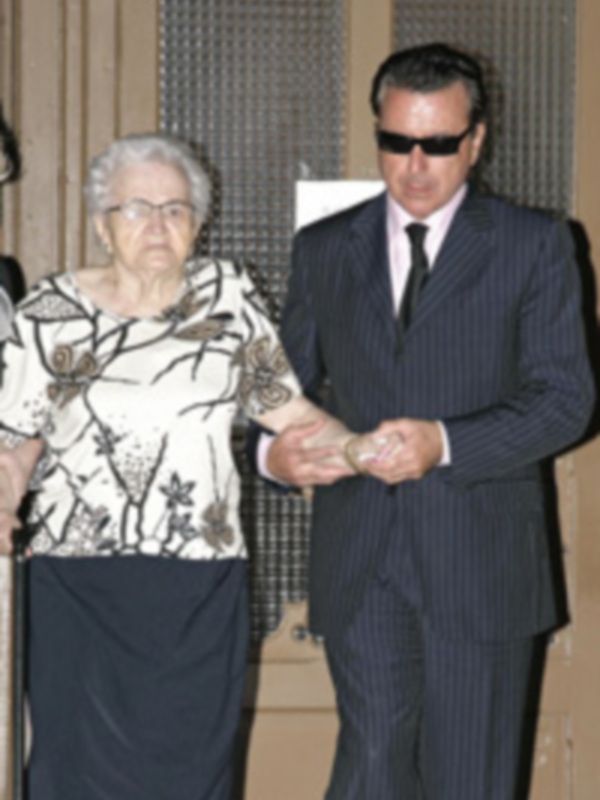 Un hombre y una mujer se saludan con una mano, ambos vestidos formalmente. El hombre lleva un traje de pana y gafas de sol, mientras que la mujer viste una blusa con estampado floral y un vestido oscuro. El fondo es neutro con una textura de madera.