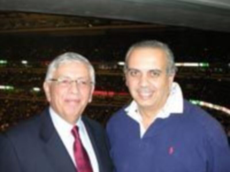Dos hombres posan juntos en un estadio, uno con traje y corbata, el otro con chaqueta azul y bufanda, ambos sonrientes y mirando hacia la cámara.